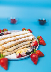 Close-up of focused French Crepes with gem and chocolate creme/sauce and icing sugar on the top and berries on the side on a blue background.