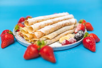 Close-up of a beautifully decorated serving plate full with French Crepes with strawberries around the plate and series inside. Homemade French crepes on blue background with free space for decoration