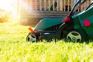 Green electric lawn mower on a freshly mown lawn in the garden against the background of a village house with flare light