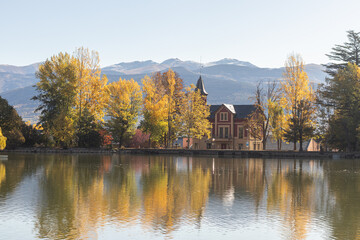 Views from a lake and beautiful trees, yellow and greens colors, good weather and sun. With a castle on the right side and sun at the end of the day.