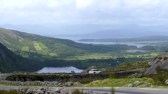 High angle scenery view from Healy Pass in Ireland. White car is driving on the road R574 above Glanmore lake and Kenmare Bay in the background; handheld camera shot.