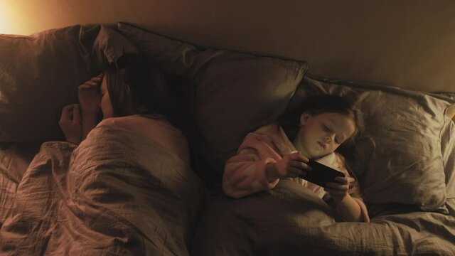 Child insomnia. Night entertainment. Girl watching video on phone in bed with sleeping older sister.