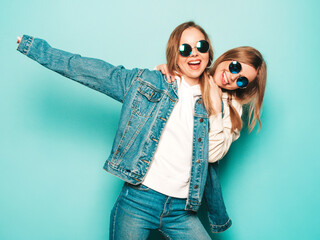 Two young beautiful brunette smiling hipster girls in trendy summer hoodie and jeans jacket clothes. Sexy carefree women posing near blue wall. Trendy and positive models having fun in sunglasses