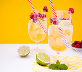 Two glasses of sparkling water, raspberries and lime slices stand on a white table against the background of an yellow wall. Near is a bowl with ripe raspberries and mint leaves, lime slices.