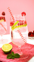 Two glasses with a drink of lime and raspberry stand on a white table against the background of a pink wall. Glasses decorated with raspberries. Near are fresh mint leaves and lime wedges. Vertically