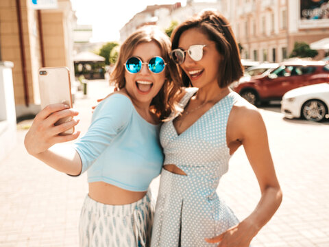 Two young beautiful smiling hipster girls in trendy summer clothes.Sexy carefree women posing on the street background in sunglasses. They taking selfie self portrait photos on smartphone