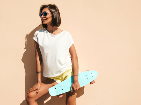 Young beautiful sexy smiling hipster woman in sunglasses.Trendy girl in summer T-shirt and shorts.Positive female with blue penny skateboard posing in the street near wall