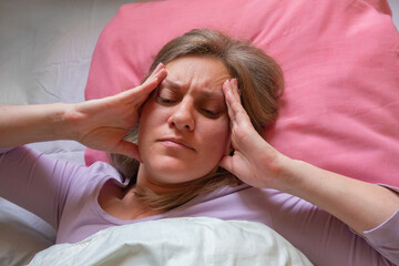 Middle aged woman has a headache. 40 years women lying in the bed and touches the head with hands. She is unwell and sick. Girl close-up portrait with health problems