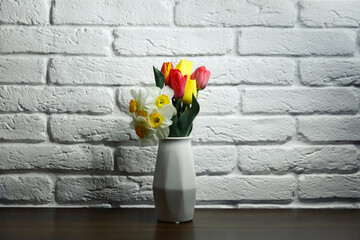 Red and yellow tulips in a white geometric ceramic vase stand on a wooden table near white brick wall. Flowers in a vase. mother's day.