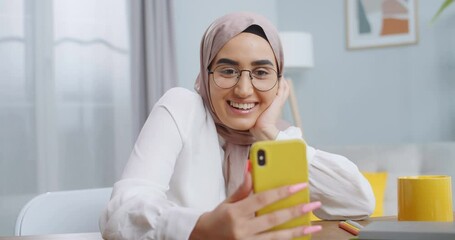 Close up of young muslim woman wearing hijab and glasses having video call using camera on yellow smartphone while sitting at cozy stylish modern home office. Young asian student speaking with family. - Powered by Adobe