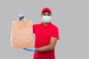 Delivery Man With Paper Bag in Hands WEaring Medical Mask and Gloves Isolated. Red Uniform Indian Delivery Boy. Home Food Delivery. Paper Bag