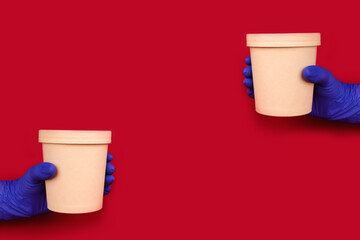 Cmposition with hands wearing blue disposable protective gloves on red background holding round paper food container - cardboard cup for soup or other dishes takeaway. Copyspace and mockup