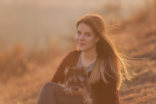 Beautiful Young Girl With Long Red Hair Developing In The Wind Hugs A Black Schnauzer Dog. She Looks At The Camera And Smiles In The Sunset. Pet Care And Travel. Dog Is Man’s Best Friend. Dog Walking