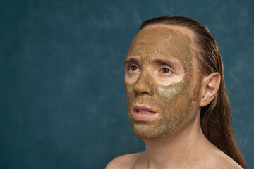 Attentively looking man with wet long hair in green clay mask on his face waiting of spa procedures.