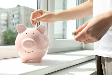 Woman putting money into piggy bank at window sill indoors, closeup