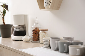 Modern coffeemaker and cups on commode near light wall indoors