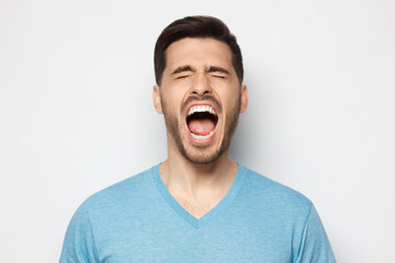 Young man in blue t-shirt screaming loudly with closed eyes, feeling strong anger and despair, suffering from trouble and exhaustion, isolated on gray background