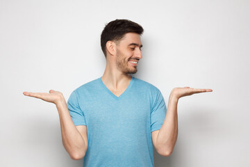 Young handsome man choosing between two different options, holding 2 hands, isolated on gray background