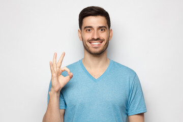 Young smiling man in blue tshirt showing OK sign or okay gesture with his fingers, isolated on gray...
