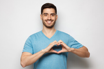 Portrait of cheerful young man in blue t-shirt showing heart sign isolated on gray background