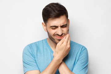 Toothache concept. Young man feeling pain, holding his cheek with hand, suffering with painful expression, isolated ion gray background