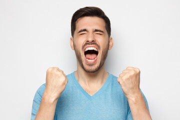 Close up of emotional man screaming with joy and closed eyes, holding hands in gesture of winner, isolated on gray background