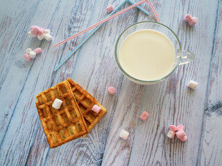 Breakfast for the child with milk, cookies and sweets. On a wooden surface is a cup of milk. Rectangular cookies, sweets and tubule for drinks on the table.