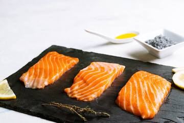 side view of three pieces of raw salmon on a slate board and lemon slices with olive oil and black salt