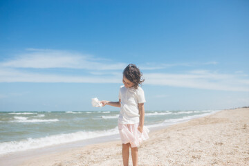 Little girl wearing light t-short and light pink skirt at the sea beach with a big beautiful seashell. Summer vacation