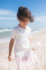 Little girl wearing light t-short and light pink skirt at the sea beach with a big beautiful seashell. Summer vacation