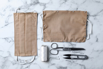 Homemade protective masks and sewing accessories on white marble background, flat lay
