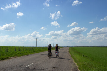 Two cyclists husband and wife village spouses ride bicycles along a rural asphalt road along a field in summer. Blue sky and white clouds.