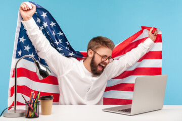 happy businessman raising American flag, watching live patriotic program on laptop at workplace and...