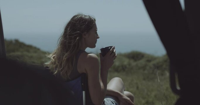 Young adult female on road trip drinking coffee outside Campervan looking at the sea