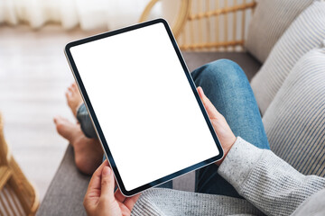 Fototapeta Mockup image of a woman holding black tablet pc with blank desktop white screen while lying on a sofa at home obraz