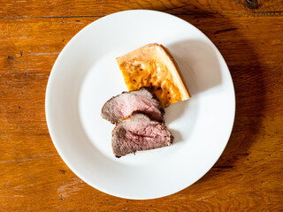 top view of home-made sliced Roast Beef and piece of Yorkshire pudding on white plate on old wooden table at home kitchen