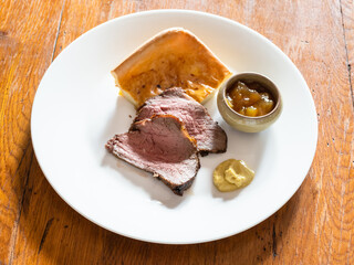 portion of home-made sliced Roast Beef and piece of Yorkshire pudding with mustard and sauce on white plate on old wooden table at home kitchen