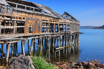 Old abandoned wooden dock reflected in calm blue water of Barents sea, rocks. Beautiful northern...