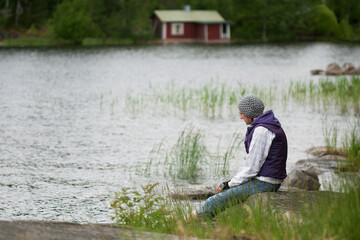 Fototapeta na wymiar Woman in hat and purple waistcoat relaxing on lakeside in breezy summer day. Woman is spending time alone to find harmony and balance. New vacation approach - responsible travel. Solo travell concept.