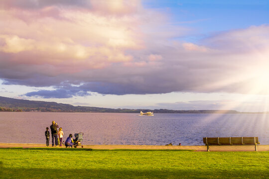 Young Family Watching A Seaplane Speeding Up To Take Off From The Lake Rotorua At Sunset