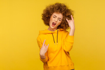 Yeah, that`s wonderful! Portrait of overjoyed curly-haired woman in urban style hoodie showing rock and roll hand sign, screaming and gesturing to heavy metal, rock music. indoor studio shot isolated