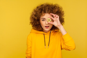 Digital money, cryptocurrency. Portrait of curly-haired woman in urban style hoodie covering eye with golden bitcoin, looking at camera through btc coin. studio shot isolated on yellow background