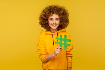 Internet trends. Portrait of happy curly-haired woman in urban style hoodie showing big hashtag sign, meaning blog recommendation, tagging viral post. indoor studio shot isolated on yellow background - obrazy, fototapety, plakaty