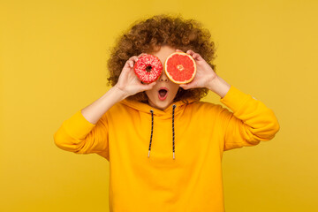 Fruits vs sugary desserts. Portrait of surprised curly-haired woman in urban style hoodie covering eyes with fresh grapefruit and sweet doughnut, open mouth in amazement. indoor studio shot isolated