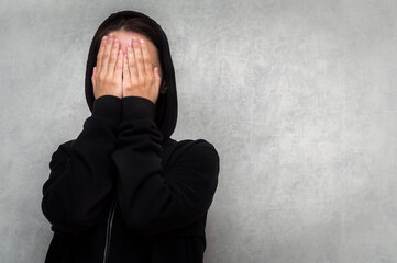 young woman in a black sweater and a hood on her head covers her face with her hands. The concept...