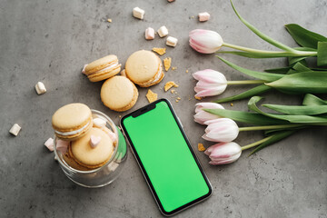 Obraz na płótnie Canvas Flat Lay Background of Fresh Tulips, Yellow French Macarons and Smartphone with Green Screen on the Table, Copy Space