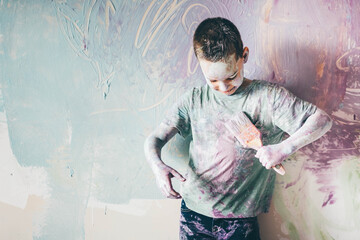 Joyful boy colours himself with wooden brush and smiles standing near purple wall.