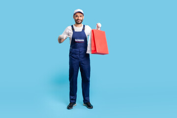 Full length happy professional courier in uniform pointing at shopping bags and smiling to camera, delivering parcel with goods ordered online at fashion store. studio shot isolated on blue background