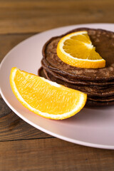 Delicious chocolate pancakes with slices of orange for breakfast on a pink plate on wooden table. Fresh, sweet and healthy dessert. Vertical