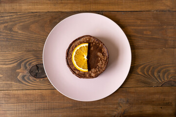 Delicious chocolate pancakes with slices of orange for breakfast on a pink plate on wooden background. Fresh, sweet and healthy dessert. Top view